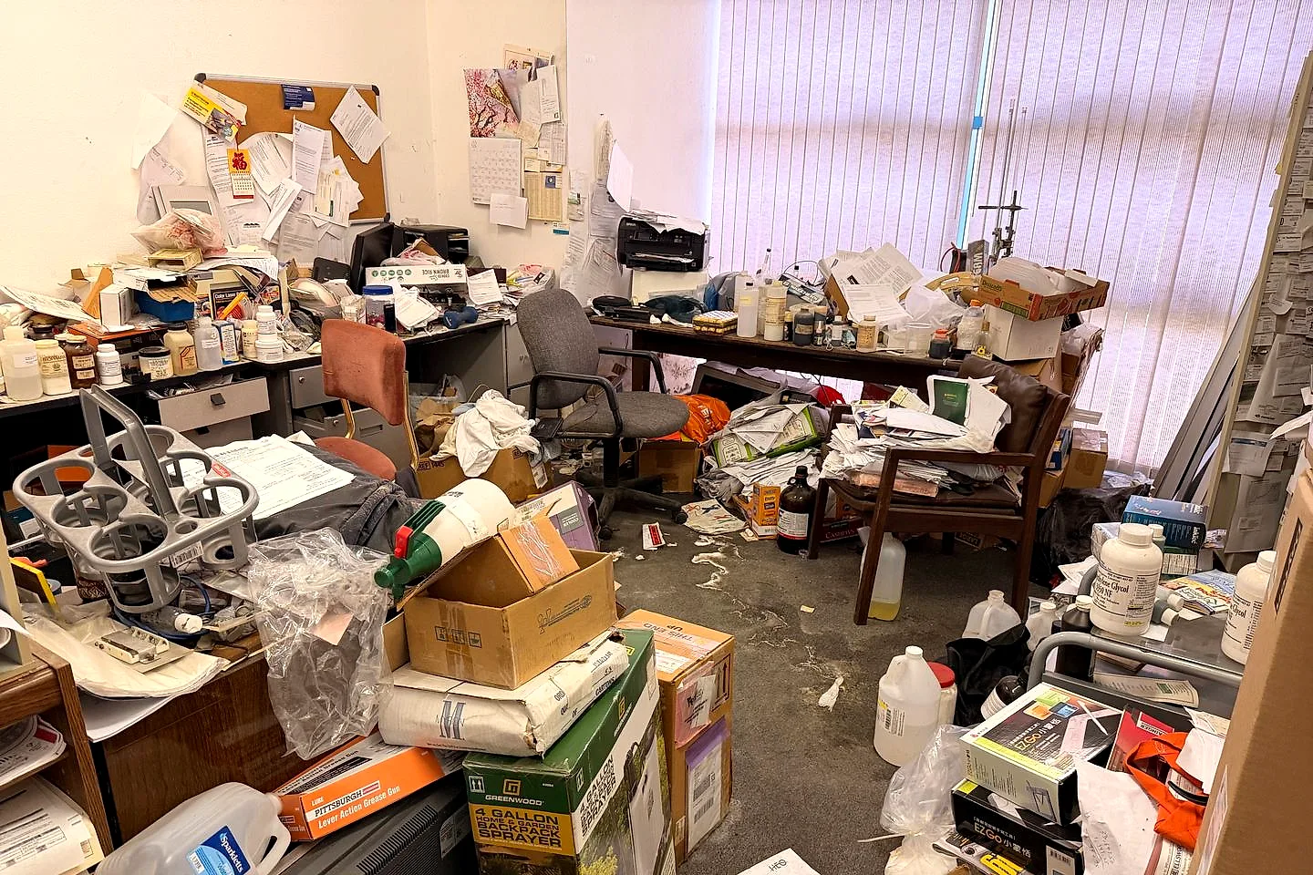 Cluttered laboratory office with bottles, jars, boxes, papers, and equipment covering desks and the floor before the lab pack